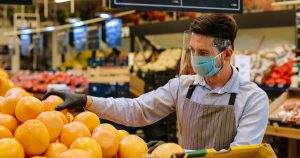 Portrait of male food store employee in face protection shield and apron standing in store and sorting fruits. Handsome Caucasian man seller in mask working indoor. Supermarket concept